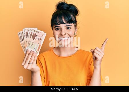 La giovane ragazza ispanica che tiene 5000 coreani del sud ha vinto le banconote sorridendo felice di puntare con mano e dito a lato Foto Stock