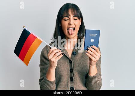 Giovane donna ispanica che tiene la bandiera della germania e passaporto che stacca la lingua fuori felice con l'espressione divertente. Foto Stock