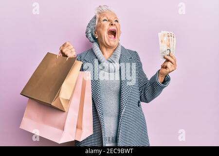 Donna anziana con capelli grigi che tiene borse per la spesa e banconote inglesi in sterline arrabbiate e pazza urlando frustrato e furioso, gridando con rabbia guardando in su. Foto Stock