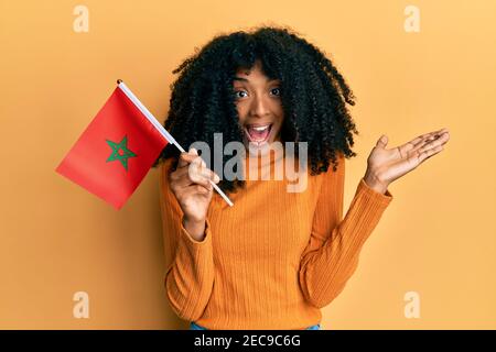 Donna afroamericana con capelli afro che tiene bandiera del marocco festeggiando successo con sorriso felice e espressione vincente con mano sollevata Foto Stock