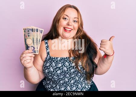 Più taglia giovane donna caucasica che tiene le banconote di yen giapponesi che indicano pollice fino al lato sorridente felice con bocca aperta Foto Stock