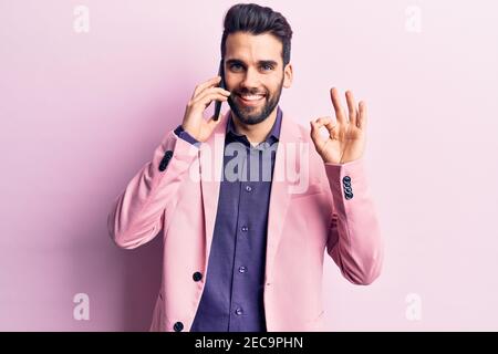 Giovane bell'uomo con barba che parla sullo smartphone facendo ok segno con le dita, sorridendo amichevole gesturing simbolo eccellente Foto Stock