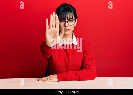 Giovane donna bruna con pangs che indossa occhiali seduti sul tavolo facendo smettere di cantare con il palmo della mano. Avvertimento espressione con negativo e serio Foto Stock