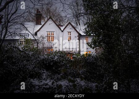 Una casa d'arte e artigianato con finestre illuminate, sparato attraverso alberi di bosco in inverno, neve a terra, Hampstead Garden sobborgo, Barnet, Regno Unito Foto Stock