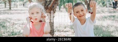 Bambini caucasici, ragazza e ragazzo che mangiano bacche in fattoria. Felici amici contadini raccolgono la raccolta stagionale di ciliegi in campagna. Estate divertente per i bambini Foto Stock