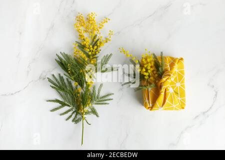 Vista dall'alto del regalo confezionato in tessuto di lino in stile furoshiki eco con fiore mimosa giallo. Concetto di celebrazione del giorno della donna o della madre. Foto Stock