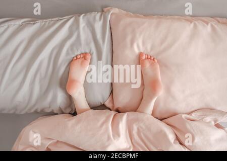 Bambini gambe gambe piedi stesi su cuscini nel letto a casa. Bambini che giocano nascondono e cercano il gioco sotto coperta. Tacchi per bambini graziosi e rosa capovolti a letto Foto Stock