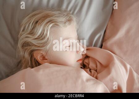 Closeup di carino caucasico bionda bambina che dorme sognando in letto a casa. Dormi un bambino stanco con gli occhi chiusi sotto la coperta rosa in camera da letto. Mattina Foto Stock