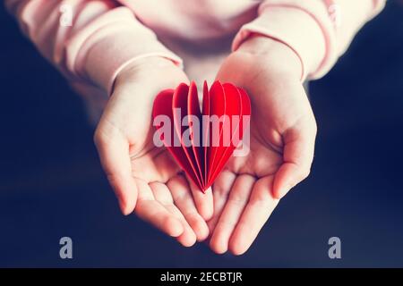 Closeup delle mani del bambino capretto che tengono carta rossa piegata cuore sfondo scuro. Festa di San Valentino. Concetto di supporto, cura e amore. Simbolo del cuore rosso di Foto Stock