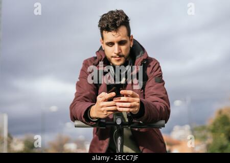 Bel ragazzo che invia un messaggio con il suo cellulare sullo scooter elettrico. Mettere a fuoco sul telefono. Foto Stock