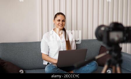 Sorridente blogger femmina registra un nuovo video. Si siede sul divano a casa e tiene un computer portatile. Concetto positivo del blog di affari. Foto Stock