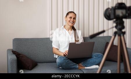 Sorridente blogger femmina registra un nuovo video. Si siede sul divano a casa e tiene un computer portatile. Concetto positivo del blog di affari. Foto Stock