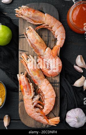 Set di gamberi o gamberetti grezzi, su tagliere di legno, su sfondo di tavola di legno nero, vista dall'alto piatto Foto Stock