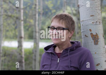 Una donna di mezza età che indossa occhiali e una camicia viola sorride mentre si appoggia contro un albero di aspen. Foto Stock