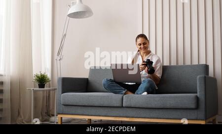 Felice giovane donna con fotocamera che usa il laptop a casa. Il fotografo guarda le foto scattate e si siede sul divano. Foto Stock