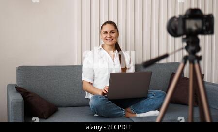 Sorridente blogger femmina registra un nuovo video. Si siede sul divano a casa e tiene un computer portatile. Concetto positivo del blog di affari. Foto Stock