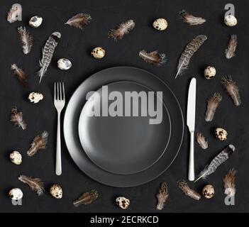 Tavolo con un piatto nero, uova di quaglia e piume su sfondo nero. Concetto di Pasqua. Vista dall'alto. Foto Stock