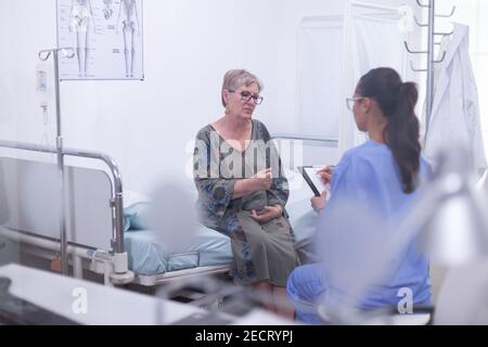 Infermiere prendendo appunti sulla clipboard consultando la vecchia donna, facendo domande a pazienti maturi riempire la forma di storia della malattia, prescrivere il trattamento. Anziana donna che parla con giovane assistente, concetto di assistenza sanitaria Foto Stock