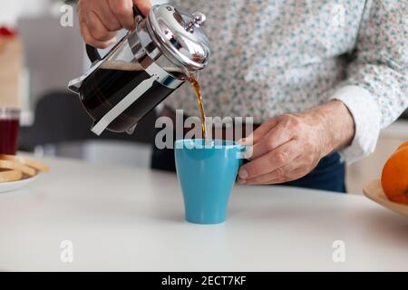 Uomo anziano che usa la pressa francese per la preparazione del caffè e lo versa in tazza. La persona anziana al mattino gusterai una tazza di caffè espresso marrone fresco con caffeina da tazza d'epoca, un filtro per rinfrescare il relax Foto Stock