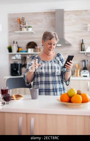 Donna anziana entusiasta che parla con la webcam del telefono cellulare con la famiglia seduta in cucina durante la colazione e sorridente. Persona anziana autentica che utilizza la moderna tecnologia Internet per smartphone. Foto Stock