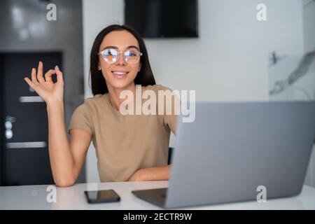 Giovane donna che usa il computer portatile in cucina facendo ok segno con le dita, simbolo eccellente Foto Stock