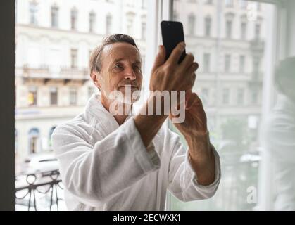 Uomo anziano che indossa accappatoio in piedi sul balcone di un hotel stanza e prendere un selfie Foto Stock