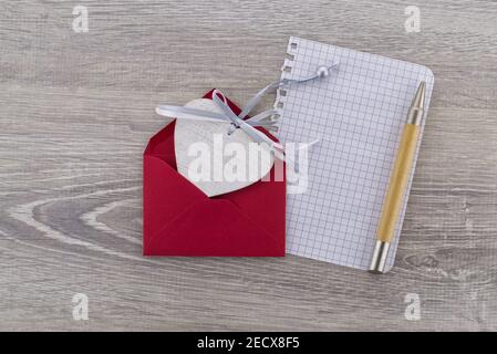 Medaglione di legno a forma di cuore posto in una busta rossa accanto a. un foglio di carta con una penna Foto Stock