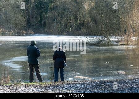 Coppia senior accanto al laghetto di pesca congelato chiamato Wilks Water vicino al canale Basingstoke a Odiham, Hampshire, Regno Unito, durante l'inverno o febbraio Foto Stock