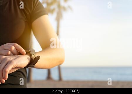 Giovane donna fitness che usa un orologio intelligente prima o dopo la corsa sullo sfondo della spiaggia al tramonto con palme. Concetto di stile di vita sano con spazio di copia Foto Stock