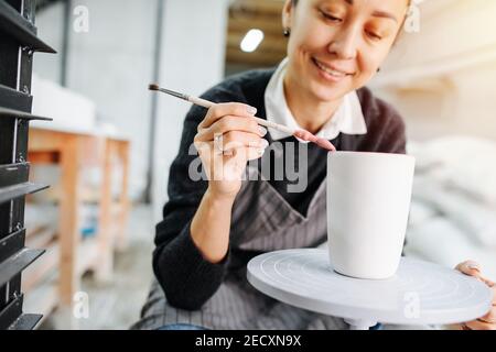 Felice lavoratrice che dipinge con cura la tazza vuota sulla ruota dei vasai in un'officina Foto Stock