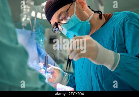 I chirurghi della squadra stanno effettuando un'operazione, il medico di mezza età bello sta guardando la macchina fotografica, in una stanza operativa moderna Foto Stock