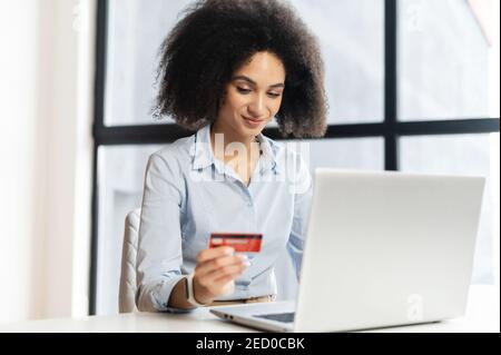 Giovane imprenditore misto-corsa seduto alla scrivania di fronte al portatile, tenendo in mano la carta di credito, inserendo il numero di carta regalo, sensazione di entusiasmo per l'acquisto Foto Stock
