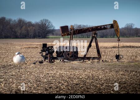 Un vecchio martinetto a pompa dell'olio arrugginito con attrezzatura di supporto si trova nel campo di mais raccolto nel Midwest americano dove gli agricoltori possono avere pompe sulla loro proprietà. Foto Stock