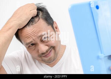 Un uomo di mezza età splende nello specchio e si preoccupa di perdita di capelli o grigio dei capelli isolato uno sfondo bianco, concetto di assistenza sanitaria Foto Stock