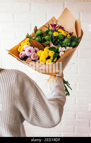 Crop persona irriconoscibile in piedi con mazzo di fiori freschi colorati sullo sfondo di muro di mattoni bianchi Foto Stock