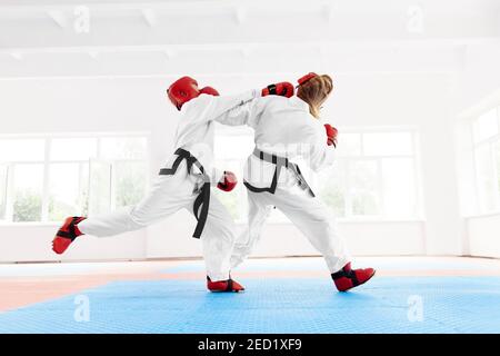 Calzare il karate femminile peoffesional indossando il kimono con la cintura nera che prende il calci alla testa dell'altro pugile. Combattente che indossa guanti di protezione rossi e casco w Foto Stock