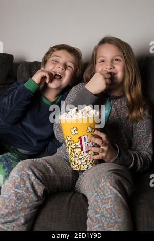 Giovani fratelli allegri bambini seduti comodi sul divano con popcorn Secchio mentre mangiano e guardano la TV Foto Stock