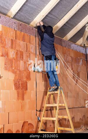 Elettricista in piedi su scala che installa i cavi nel canale / scanalatura insieme alle scatole elettriche in casa non finita costruita di mattoni di blocco di argilla. W Foto Stock