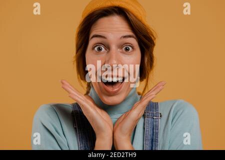 Primo piano foto di carino agricoltore femminile sorpreso di qualcosa. Indossa tute e cappello in denim, fondo isolato di colore marrone Foto Stock