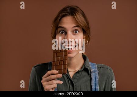 Foto di donna contadina carina, mangia cioccolato fondente con piacere. La donna indossa tute in denim, fondo isolato di colore marrone Foto Stock