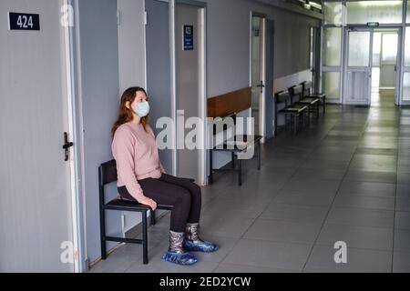 Donna in maschera di faccia siede nel corridoio di una clinica vuota senza persone e pazienti. Concetto di problema di recupero e uscita dalla clinica Foto Stock