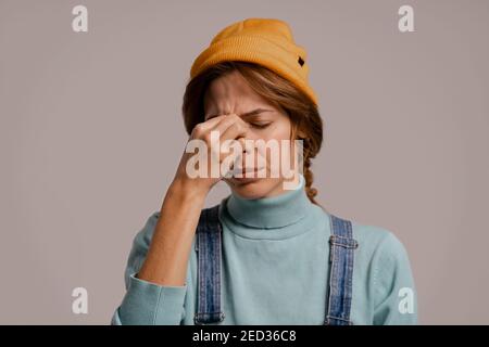 Foto di cute hipster femminile tiene la testa sembra ha mal di testa. Donna bianca indossa tuta in denim e cappello isolato di colore grigio sfondo Foto Stock