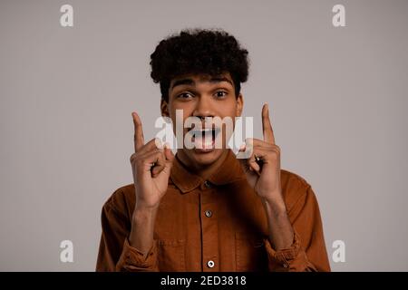 Foto di uomo nero scioccato e felicemente sorpreso di alcune notizie, punta le dita in su. Il maschio indossa una camicia, isolato sfondo grigio. Foto Stock