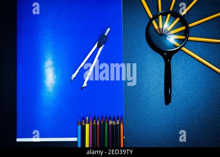 Sulla parte superiore di un grande notebook blu è posta una bussola, sul lato una lente di ingrandimento con matite, alla base dei pastelli. Foto Stock