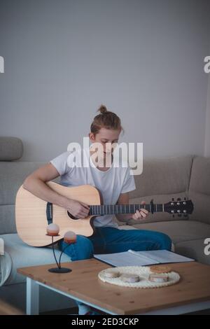 Uomo reale con capelli biondi e una T-shirt bianca si siede su un divano in una stanza illuminata, cercando di imparare accordi su una chitarra acustica di legno. Lettura delle note e. Foto Stock