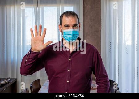 Uomo bearded in una camicia viola e una maschera per proteggersi dal coronavirus che agita con una mano. Concetto di salute Foto Stock