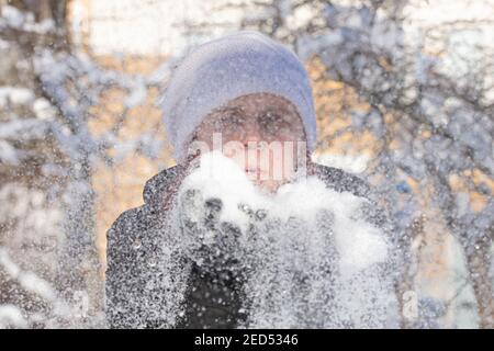 Ritratto di una donna allegra in un cappello bianco invernale sulla strada. La neve è nelle mani, soffia su di essa. Foto di alta qualità Foto Stock