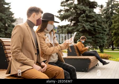 Giovane coppia in maschere protettive e casualwear elegante su cui riposarsi panca in parcheggio mentre si guarda il display del tablet e si guarda video online Foto Stock