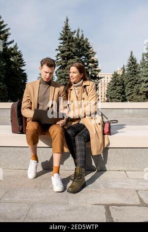 Giovane coppia affettuosa in eleganti trench beige, pullover e pantaloni seduti l'uno vicino all'altro mentre si rilassa nel parco in giornata di sole Foto Stock