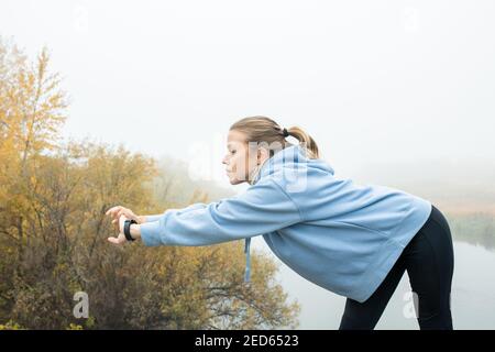 Giovane sportivo biondo con felpa con cappuccio blu e stretching leggins nero le braccia davanti a se stessa mentre si esercita in naturale ambiente Foto Stock